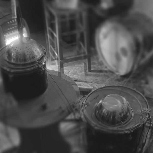 Greyscale and blurry (via double-exposure) scene of a metal bowl and colander, upturned and atop floor and rack toms. The rack tom is ontop of a small table. A bass drum, some chair legs, and general practice space clutter in the background.