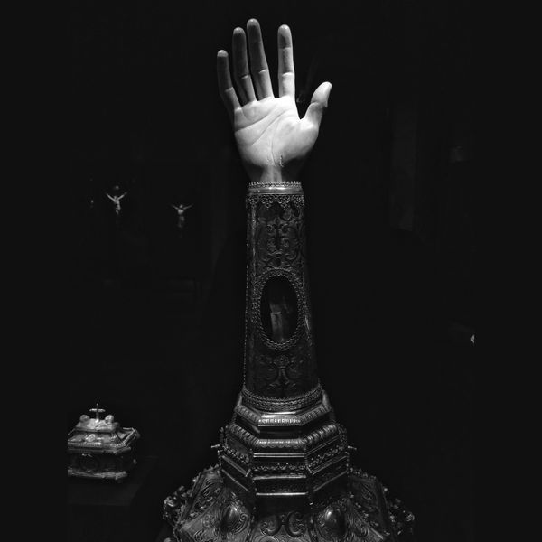 In the centre of the frame is a Christian relic; an ornate vessel, reportedly containing a bone fragment of a saint or Jesus or something (I can't remember), and topped by a hand. The image is in black and white with high contrast so the relic stands out against the blackness. Just behind, so small as to be almost indiscercnable, are two crucifixes.