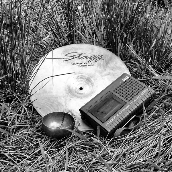 Centring the frame are a cymbal, cassette player, and singing bowl with small chain draped over its side. They are nestled amongst tall grasses. All in greyscale.