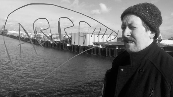 Picture of me, all hipster doofus-like with moustache and perma-beanie. I'm wrapped up warm, standing infront of water and harbour industrial buildings in Svendborg, Denmark. A badly scrawled speech bubble has been added (via phone image markup-vibes) saying 'ARRGH!!' and the whole thing is in moody greyscale to match the website aesthetic.