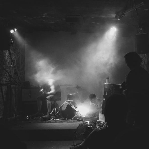 Photographed from the back of a small venue during a live performance, there are two figures obscured by smoke and stage lighting.